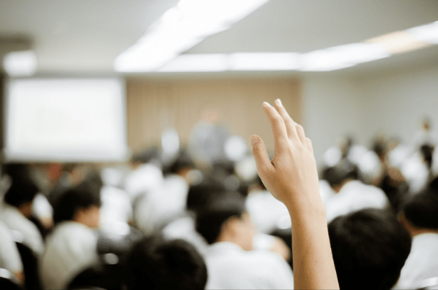 A close-up of a hand being raised in a classroom full of students